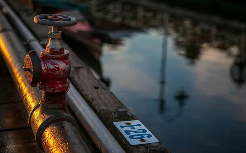 Red fire hydrant on dock with water reflection at sunset.